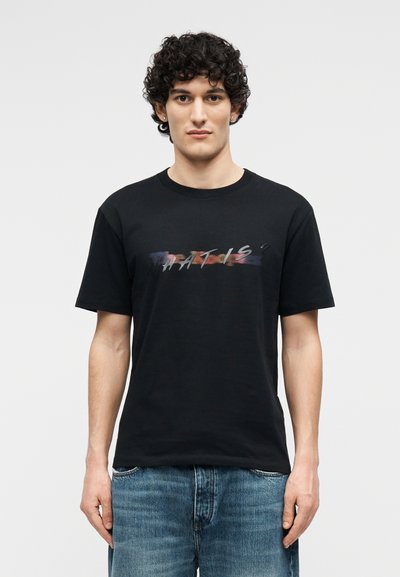 Νέος με σγουρά μαλλιά φοράει μαύρο μπλουζάκι με πολύχρωμο κείμενο και μπλε τζιν, στέκεται μπροστά σε απλό ανοιχτό φόντο.