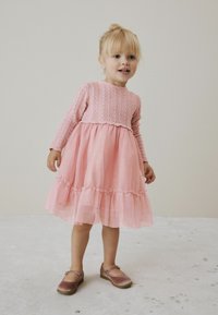 Vaaleanpunainen mekko, jossa on pitkät hihat, teksturoitu neulepäällinen, kerroksellinen tyllihelma ja kimallusta. Yhdistetty vaaleanpunaisiin kenkiin, joissa on remmi.