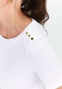 T-shirt bianco in cotone con maniche corte, caratterizzato da tre bottoni decorativi dorati sulla spalla. Texture liscia, scollatura rotonda classica.