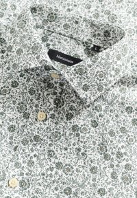 Vit skjorta med grönt blommönster, med en klassisk krage och ljusbeiga knappar. Märkesetikett "Matinique" synlig inuti kragen.