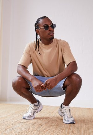 Camiseta de manga corta beige, pantalones cortos de mezclilla azul claro y zapatos deportivos grises. La persona está en cuclillas sobre una alfombra texturizada de color claro.