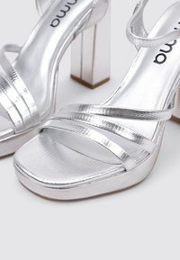 Sandales à talons plateformes en argent avec plusieurs brides texturées, orteils carrés et un grand talon en block. Finition lisse et brillante avec des détails de design.