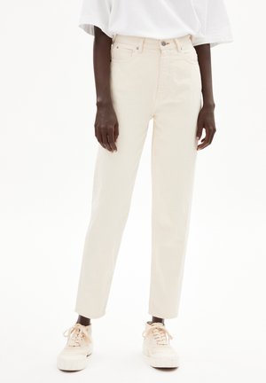 Personne portant un jean droit blanc cassé, un t-shirt blanc oversize et des baskets assorties blanc cassé, debout devant un fond uni.