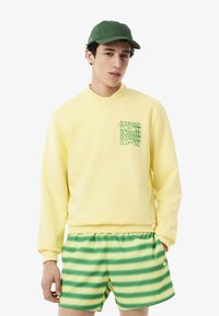 Gele sweatshirt met een groene bedrukte logo op de borst, gecombineerd met gestreepte shorts in groen en geel, en een groene pet.