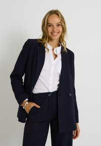 Traje de raya diplomática en color navy que presenta un blazer de doble botonadura y pantalones a juego, combinado con una camisa blanca de botones. Textura de tela suave.