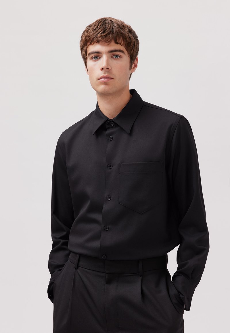 Schwarzes Langarmhemd mit Knöpfen und Kragen, einer einzelnen Brusttasche, glatter Textur und passend zu einer schwarzen Hose.