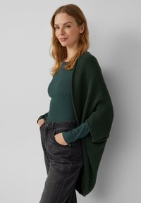 Dunkelgrüne Strickjacke über einem enganliegenden grünen Oberteil, kombiniert mit schwarzen Jeans. Die Strickjacke ist offen und hat eine strukturierte Oberfläche sowie weite Ärmel.