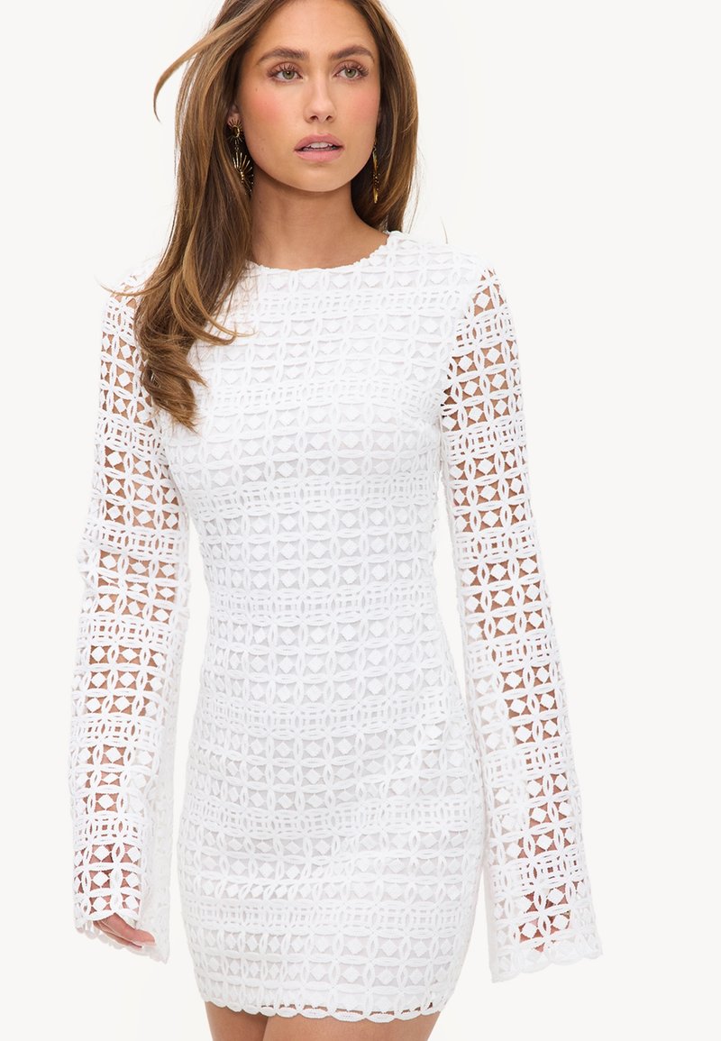 Femme portant une mini-robe blanche à manches longues avec un motif en dentelle géométrique et des bords festonnés aux manches et à l'ourlet.
