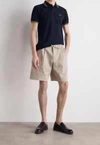 Tummansininen lyhythihainen polo-paita, jossa on keltaisia yksityiskohtia, yhdistettynä beigeihin laskostettuihin shortseihin ja mustiin loafereihin, joita käyttää seisova miesmalli.