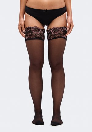 Un mannequin porte une culotte noire et des bas autofixants transparents avec des revers en dentelle à fleurs roses, sur un fond clair uni.