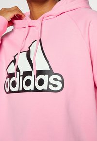 Sweat à capuche rose en mélange de coton, avec un grand logo Adidas noir sur le devant. Des cordons de serrage à la capuche complètent le design.