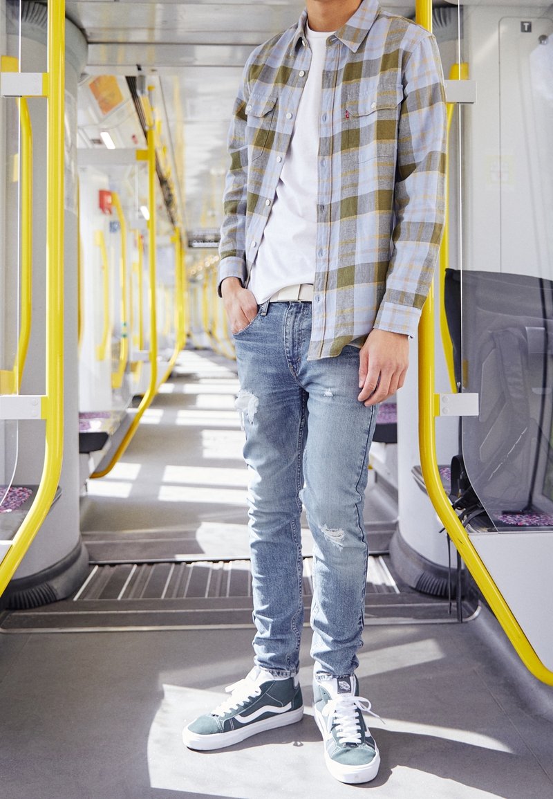 Camisa de cuadros abotonada en azul y verde, camiseta blanca, vaqueros azules desgastados y zapatillas verdes. Escena dentro de un vehículo de transporte.