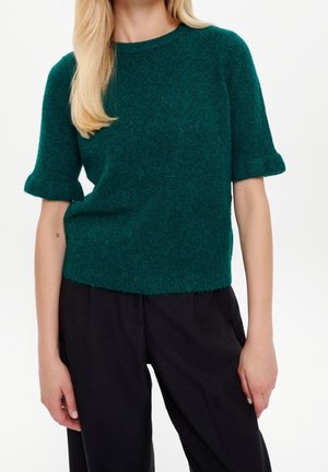 Kvinde iført en mørkegrøn kortærmet sweater og sorte bukser, stående med hænderne afslappet ned langs siden mod en hvid baggrund.