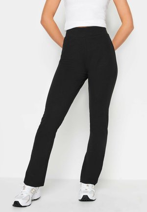 Personne portant un pantalon noir droit et des baskets blanches, debout les mains dans le dos devant un fond clair uni.