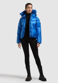 Modrá prešívaná bunda s kapucňou, vybavená dvoma vreckami na zips a lesklým povrchom. Kombinovaná s čiernymi legínami a čiernymi šnurovacími čižmami.
