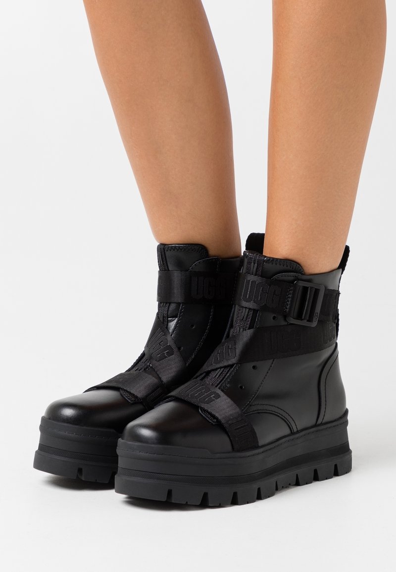 Mustat nilkkurit kiiltävällä pinnalla, joissa on paksut pohjat, säädettävät hihnat ja edessä vetoketju helppoa pukemista varten.