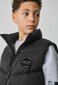Crni puffer prsluk s visokim ovratnikom, patentnim zatvaračem sprijeda i okruglom logotip oznakom s natpisom "BAKER", nošen preko svijetlosivog dukserice.