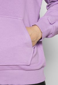 Lavendelfarbener Hoodie mit gerippten Bündchen und Saum, ausgestattet mit einer vorderen Kängurutasche und weichem Material. Nahaufnahme einer Hand in der Tasche.