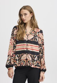 Blouse à manches longues avec un fond noir orné de motifs floraux et de motifs paisley complexes en rouge, orange et doré. Fermeture à boutons sur le devant et poignets élastiques.