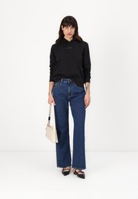 Felpa nera con cappuccio e lacci, jeans a gamba larga blu e ballerine nere. Il modello tiene una borsa a tracolla beige chiaro. Design semplice e moderno.