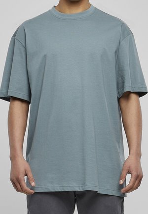 Mann trägt ein lockeres, kurzärmeliges, hellblaues T-Shirt und dunkle Hosen, steht vor einem einfarbigen grauen Hintergrund.