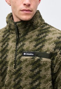 Άνδρας φορώντας πράσινο και καφέ fleece μπουφάν Columbia με σχέδιο καμουφλάζ, ψηλό γιακά και τσέπη στο στήθος.