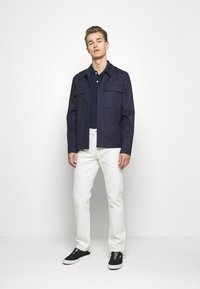 Navy button-up jacket with two chest pockets, paired with a dark polo and white jeans. Black sneakers complete the outfit.
