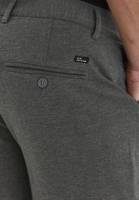 Close-up of dark gray pants pocket with button and small black label reading "ALL DAY, EVERY DAY DENIM," worn by a person with hand in pocket.
