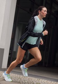 Atletyczna kobieta w miętowym topie na ramiączkach i czarnych szortach, nosząca czarną kurtkę przeciwdeszczową, biegnąca w lekkich niebieskich butach sportowych po kamiennej ścieżce.