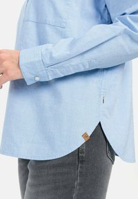 Lichtblauwe blouse met knopen en een voorzak, afgeronde onderkant en een leren logo-tag. Heeft lange mouwen met knoopmanchetten.