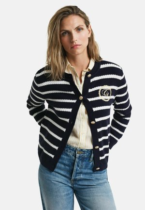 Cardigan a righe blu navy e bianche con bottoni dorati, dotato di una toppa con logo. Indossato sopra una camicia a righe chiare e jeans blu.