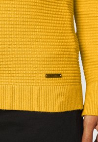 Pull en maille jaune texturé avec ourlet et poignets côtelés, étiquette en métal de la marque Esprit sur le bas devant, porté avec un pantalon noir et une main partiellement visible.