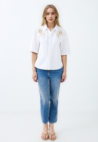 Weißes Hemd mit kurzen Ärmeln, das mit goldfarbener Blumenstickerei versehen ist. Kombiniert mit hellblauen, leicht verwaschenen Jeans und Sandalen.