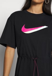 Svart T-shirt med korta ärmar, med en framträdande rosa och vit Nike swoosh-logotyp och en justerbar dragsko i midjan.