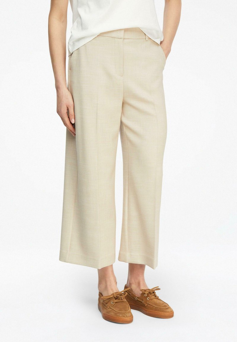 Personne portant un pantalon beige à jambes larges et raccourci, un haut blanc à manches courtes, et des chaussures bateau en suède marron à lacets, debout les mains dans les poches.