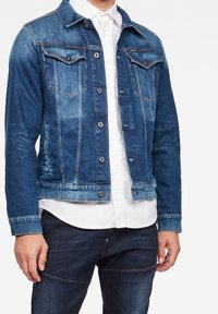 Man som bär en blå denimjacka med knappstängning över en vit skjorta och mörka jeans, stående mot en vit bakgrund.