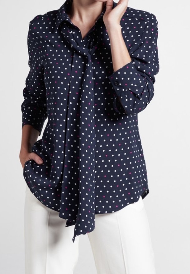 Femme portant un chemisier bleu marine à pois blancs et roses, un foulard noué autour du cou, et un pantalon blanc, posant avec une main dans la poche.