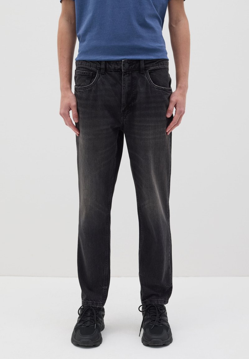 Jean en denim noir avec une coupe droite, dotés de cinq poches, d'une fermeture à bouton et d'une légère décoloration au niveau des genoux, portés avec des chaussures de sport noires.