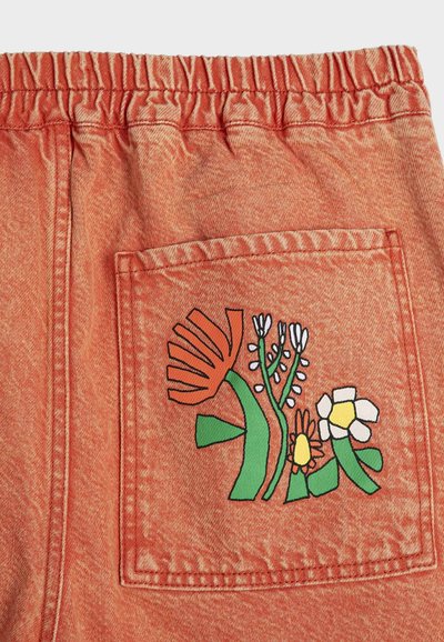 Zadný vrecko na oranžových látkových nohaviciach s elastickým pásom, zdobené farebnou vyšívanou kvetinovou potlačou v zelenej, bielej, žltej a oranžovej farbe.