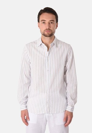 Uomo con capelli corti e scuri e barba che indossa una camicia bianca a maniche lunghe con leggere righe verticali e pantaloni bianchi, in piedi davanti a uno sfondo semplice.