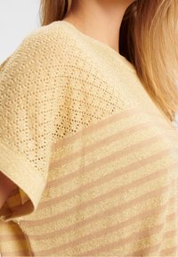 Sweter z dzianiny w jasnym żółtym kolorze z teksturowanym, perforowanym wzorem na górnej połowie i poziomymi paskami na dolnej połowie, z delikatnym połyskiem.