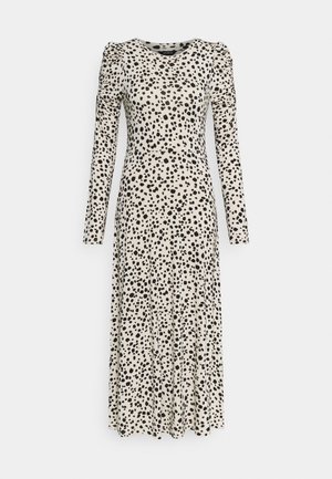 Beige väriyks pitkähiha mekko, jossa mustia epäsäännöllisiä pallokuvioita, poimutetut hartiat ja vartalonmyötäinen vyötärö, joka jatkuu liehuvaan hameeseen.