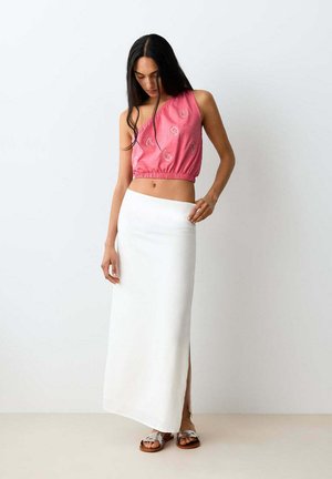 Femme portant un crop top rose à une épaule et une jupe longue blanche taille haute avec une fente latérale, debout et regardant vers le bas.