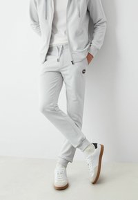 Gaiši pelēka ar rāvējslēdzēju aizdaram mīksta auduma džemperis un atbilstošas sporta bikses, kas aprīkotas ar sānu kabatām un ribotām aprocēm, kombinētas ar baltām kedām.