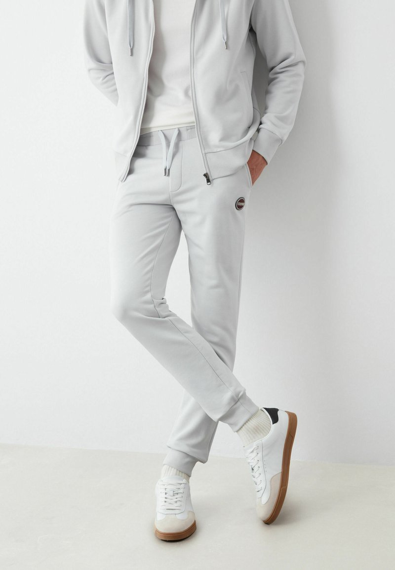 Gaiši pelēka ar rāvējslēdzēju aizdaram mīksta auduma džemperis un atbilstošas sporta bikses, kas aprīkotas ar sānu kabatām un ribotām aprocēm, kombinētas ar baltām kedām.