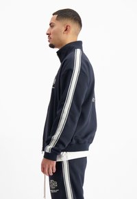 Marineblauwe ritssluitingjack met hoge kraag, witte strepen langs de mouwen en geborduurd logo op de borst, gecombineerd met bijpassende marineblauwe sweatpants.