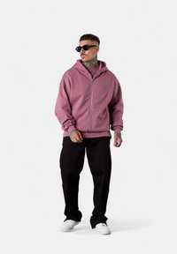 Hooded zip-up sweatshirt in soft pink, with a relaxed fit and ribbed hem. Paired with black pants and white sneakers, accessorized with sunglasses.