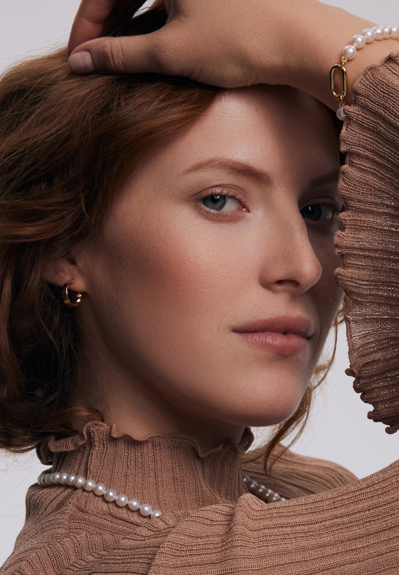Brązowy prążkowany golf z falbaniastymi akcentami, w połączeniu z perłową naszyjnikiem i złotymi kolczykami w kształcie kółek. Gładka skóra i miękkie, falowane włosy.