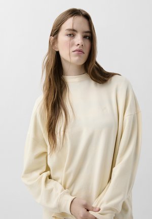 Krémový oversized svetr s vysokým výstřihem, volným střihem a žebrovanými manžetami. Hladká textura s jemným logem na hrudi.