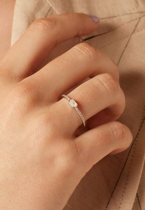 Silver ring with a hexagonal stone on a delicate band, featuring a textured pattern, worn on a hand against a neutral fabric background.